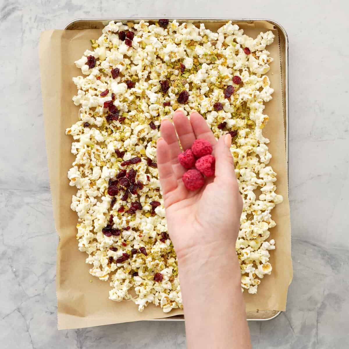 Hand holding four freeze fried raspberries above a tray of Popcorn with melted white chocolate, cranberries, pistachios on top.