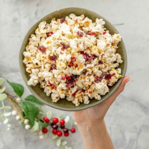 Hand holding Christmas Popcorn in a ceramic bowl above bench top with Christmas decorations around.