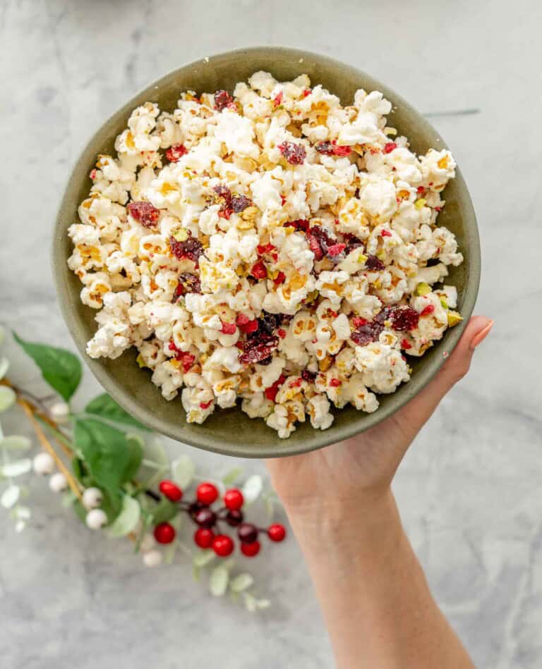 Hand holding Christmas Popcorn in a ceramic bowl above bench top with Christmas decorations around.