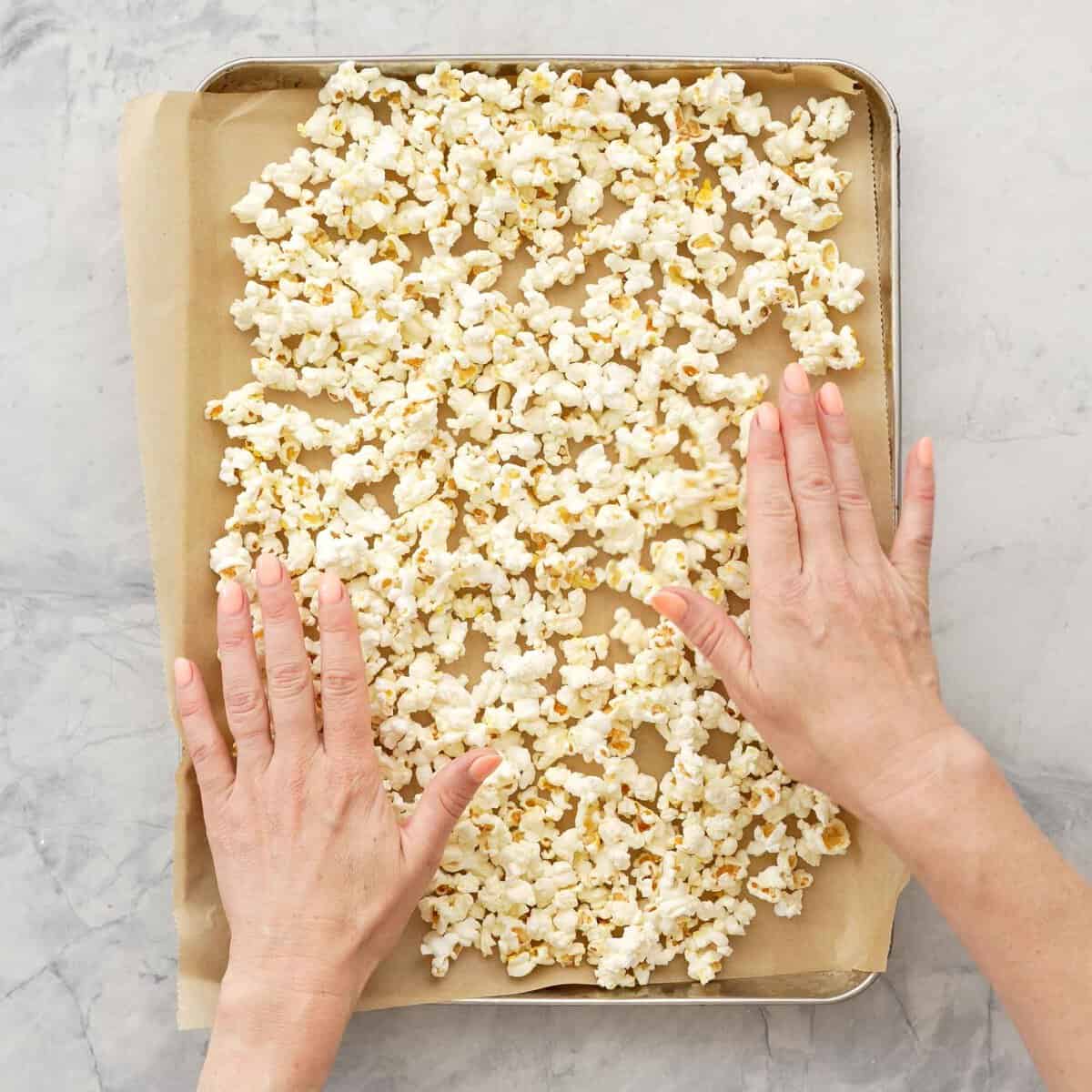 Two hands flattening out popcorn on a tray with parchment paper.