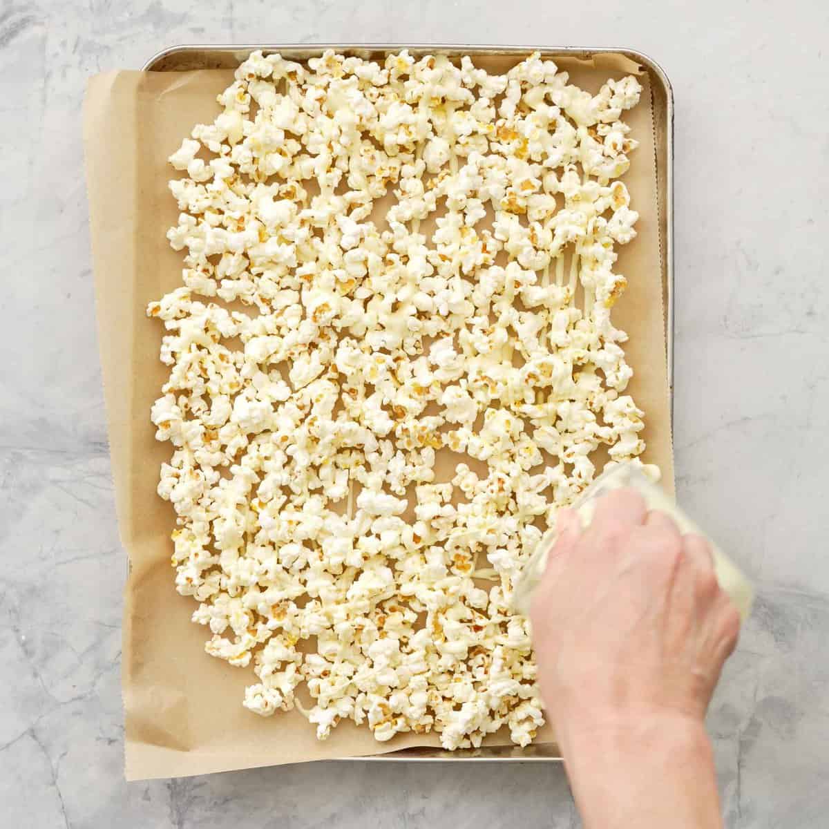 Tray of popcorn on parchment paper with hand pouring a small glass jug of melted white chocolate on top.