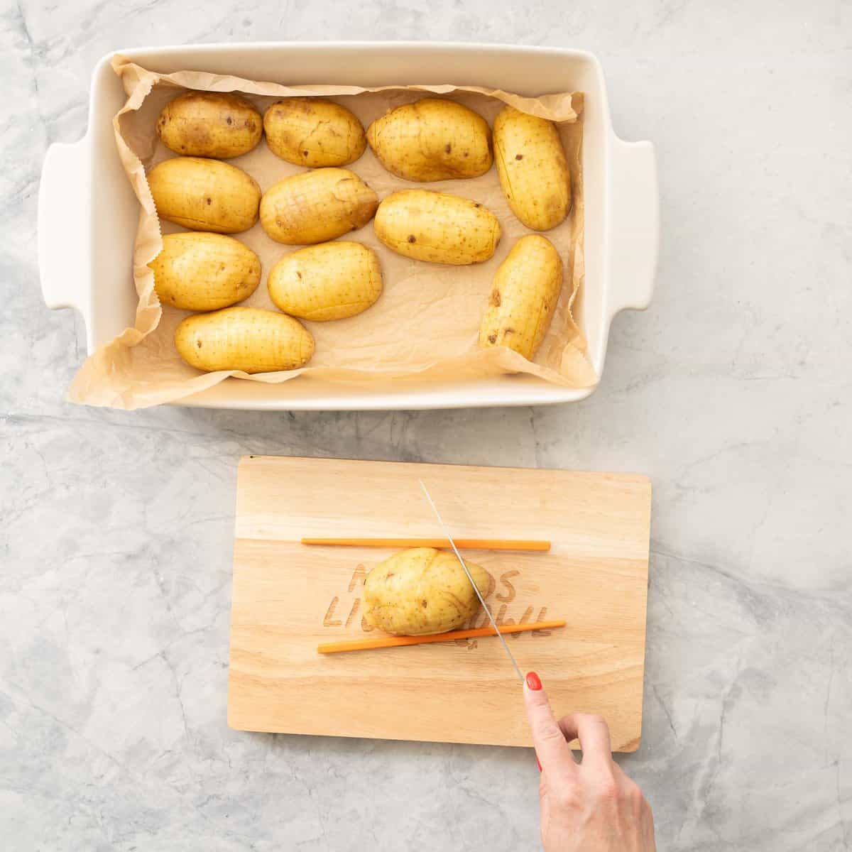 Potatoes cut in half length ways in roasting dish, one half potato on chopping board with two chopsticks on either side with knife cutting little squares into it.