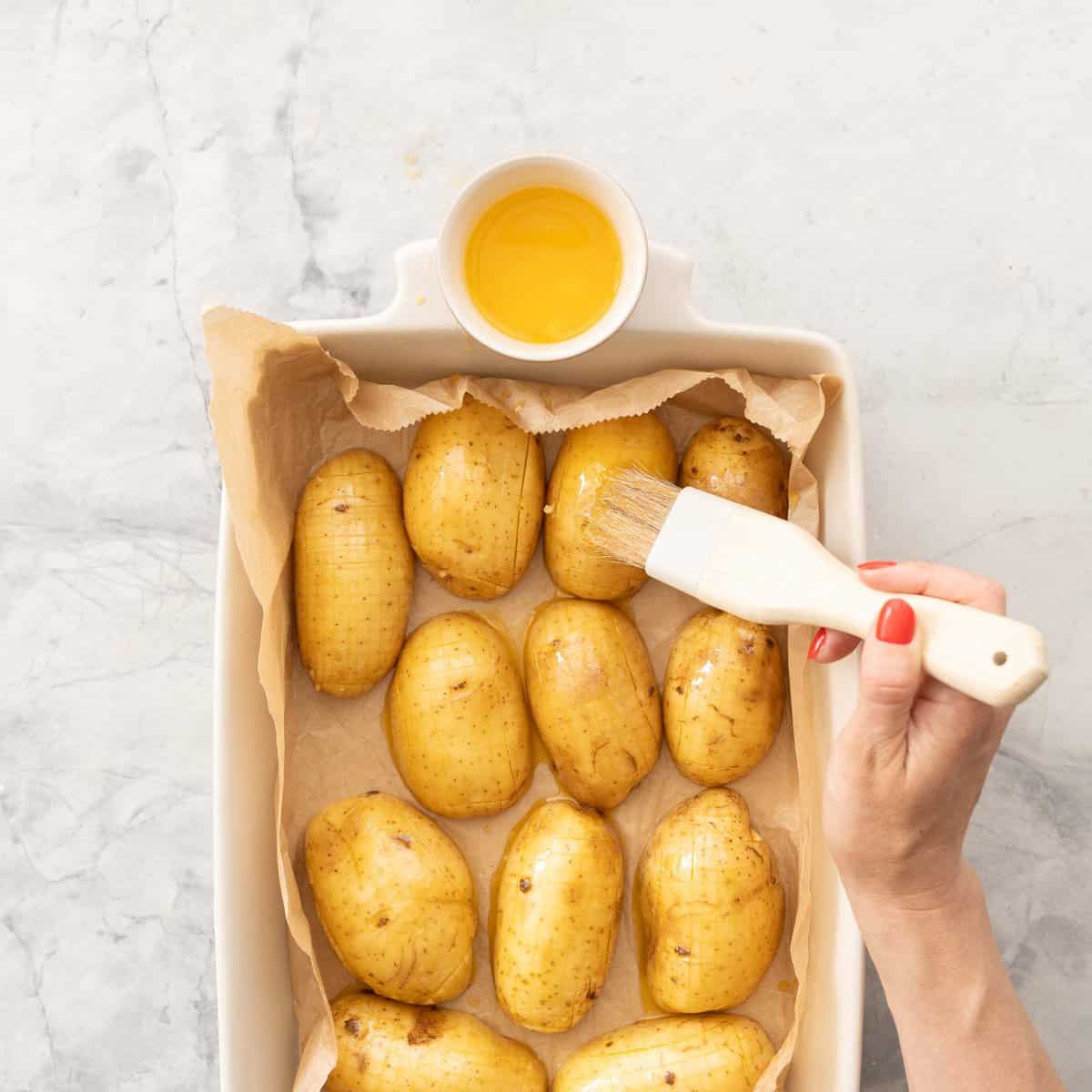 Baking dish lined with parchment paper with sliced potatoes inside, hand brushing melted garlic butter onto potatoes.