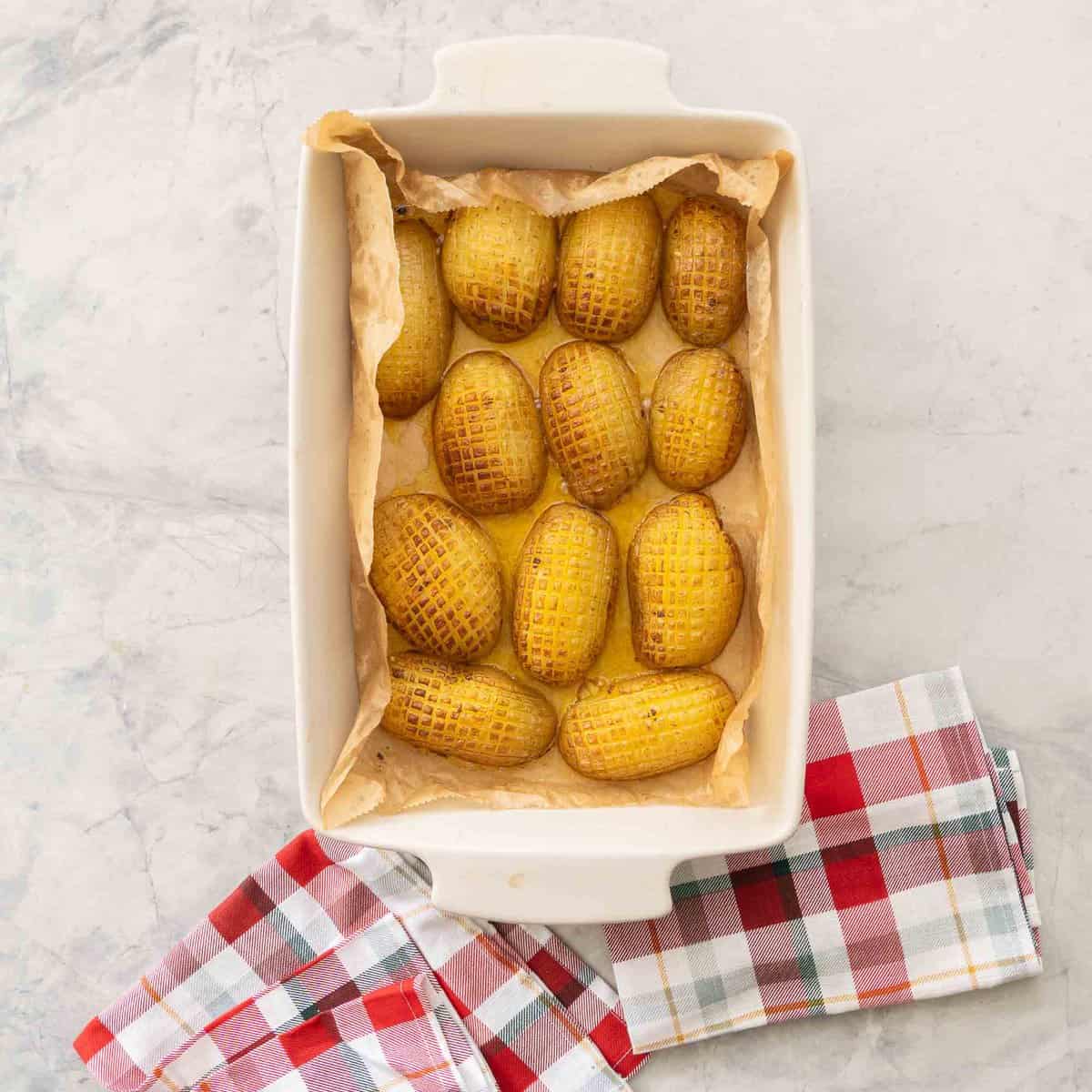 Roasted sliced potatoes in a baking dish on benchtop with christmas checkered tea towel.