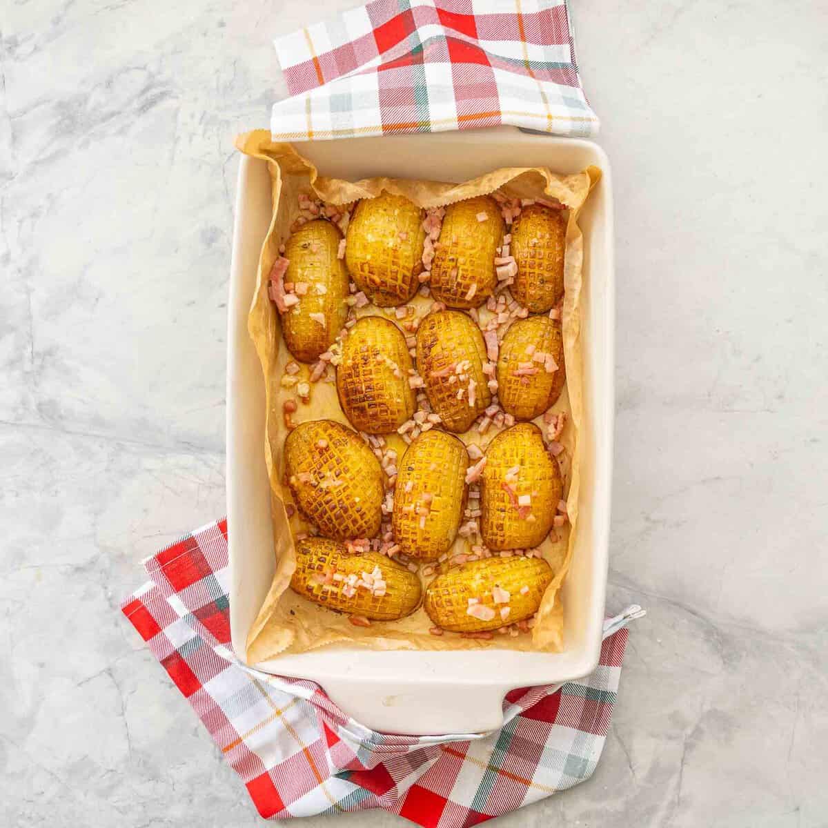 Baking dish with baked potatoes inside and diced bacon sprinkled on top on benchtop with christmas checkered tea towels.