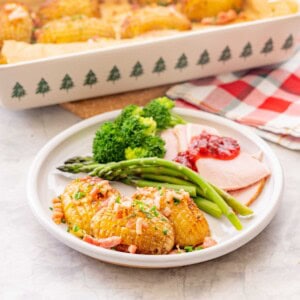 Christmas potatoes garnished with bacon and chives, served on a plate with asparagus broccoli and Christmas ham with jam on top.