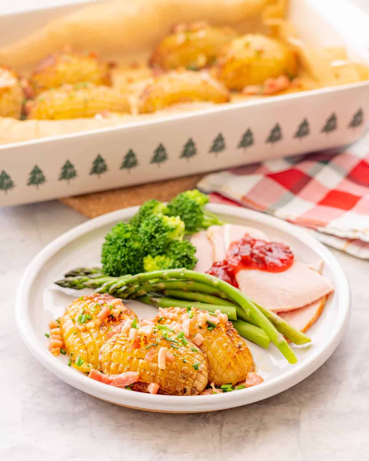 Christmas potatoes garnished with bacon and chives, served on a plate with asparagus broccoli and Christmas ham with jam on top.