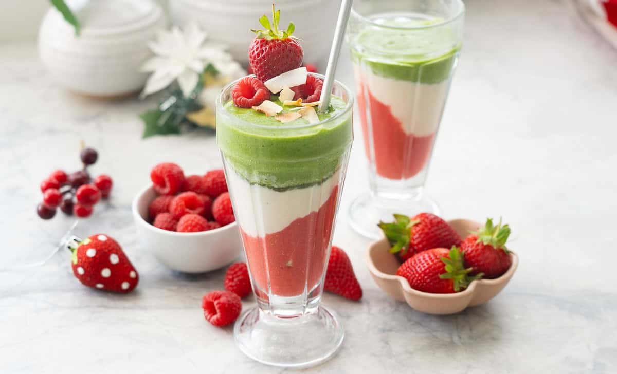 Tall cup with layered Christmas smoothies inside, red at the bottom, white in the middle and green smoothie at the top. Strawberries and raspberries on bench top and in small ramekins in background.