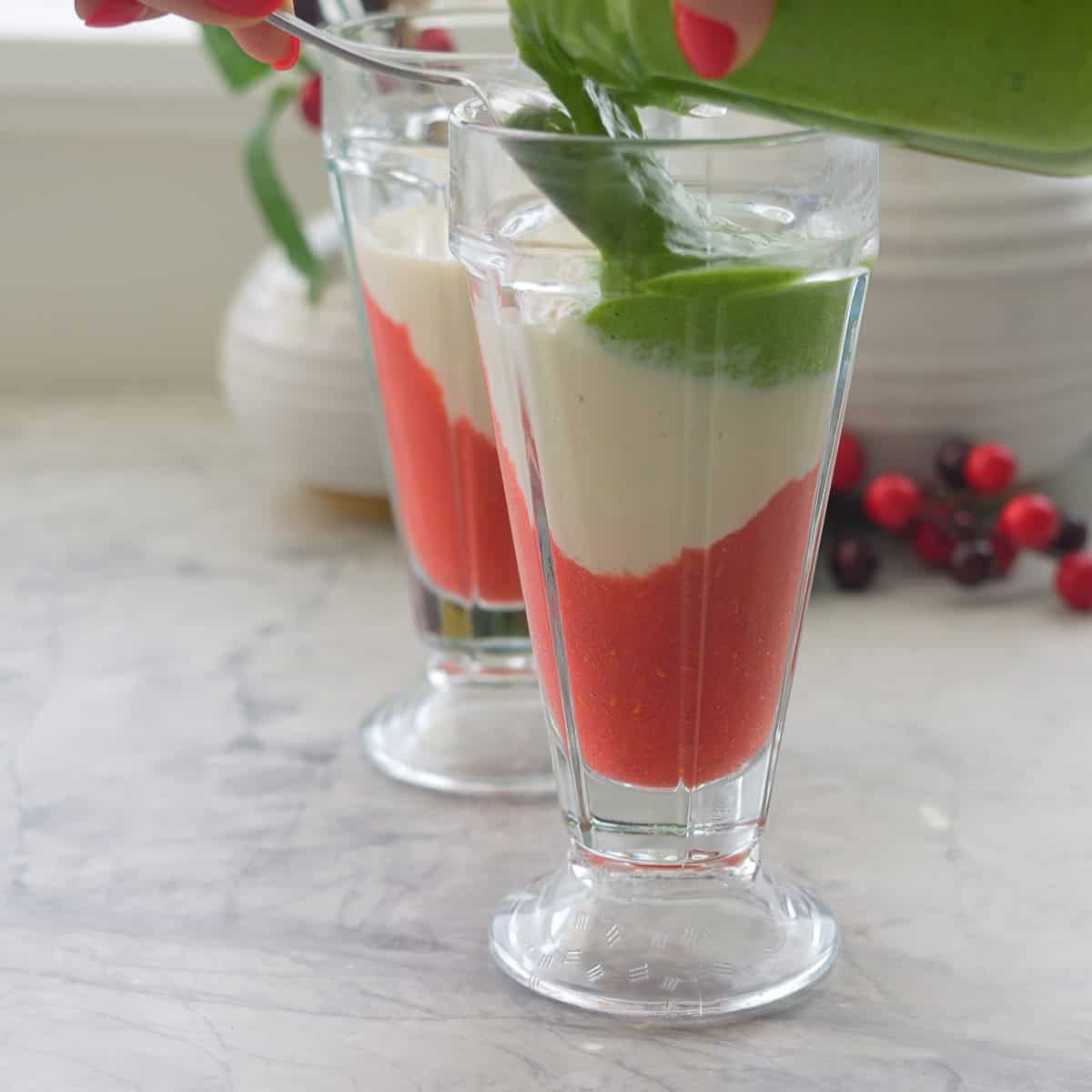 Green smoothie being poured onto red and white smoothie layers in tall glass on benchtop.