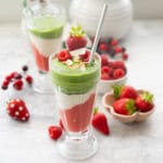 Tall cup with layered Christmas smoothies inside, red at the bottom, white in the middle and green smoothie at the top. Strawberries and raspberries on bench top and in small ramekins in background.