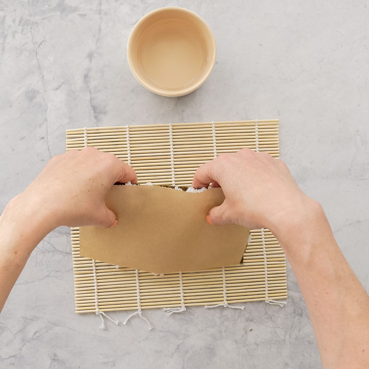 Sushi mat on bench top with small ramekin of water, Hands turning over sheet of baking paper with nori and sushi rice on top.