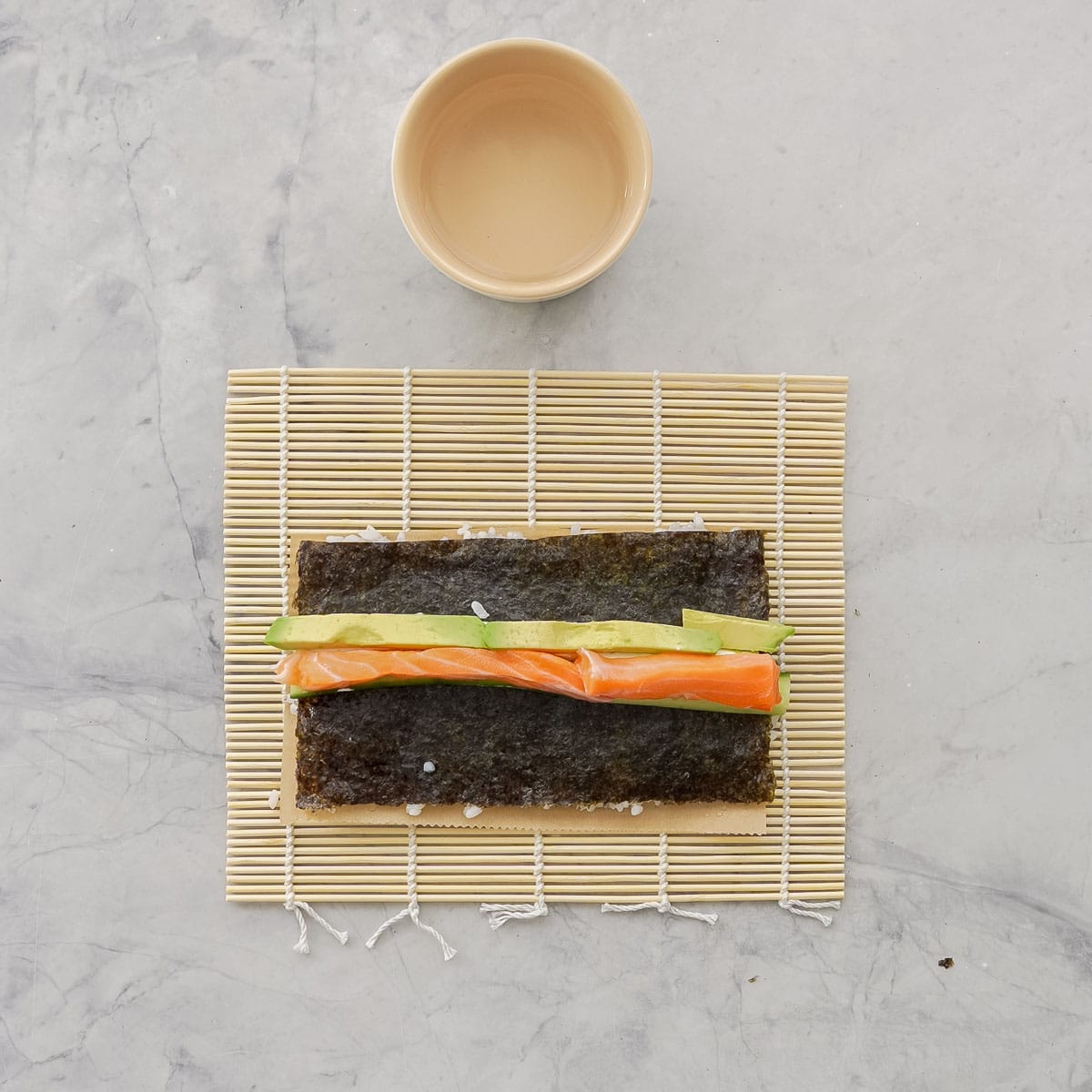 Sushi mat on bench top with baking paper on top layered with sushi rice, nori, avocado and salmon.