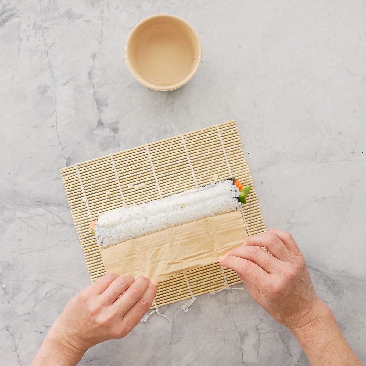 Hands holding baking paper, pulling it away from roll of sushi exposing roll of sushi and rice.