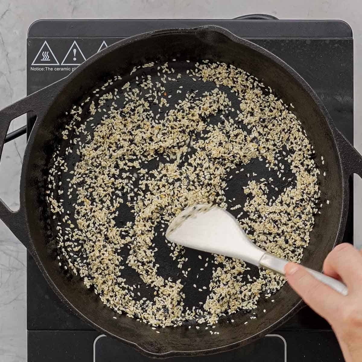 Sesame seeds on stove top in a cast iron pan being stirred with spatula.