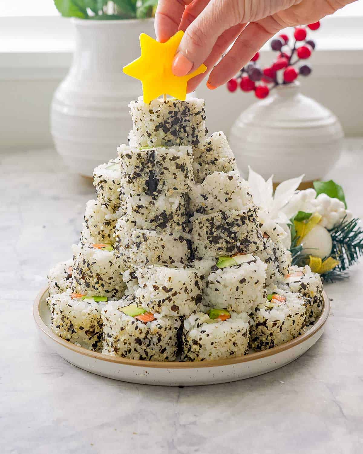 Stack of christmas sushi on a plate with hand putting a yellow capsicum star cut out with toothpick into the top of stack of sushi.