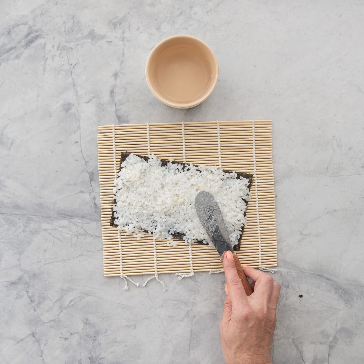 Sushi mat with nori paper on top on bench top, hand holding a metal spatula spreading sushi rice over nori, Small ramekin with water next to it.