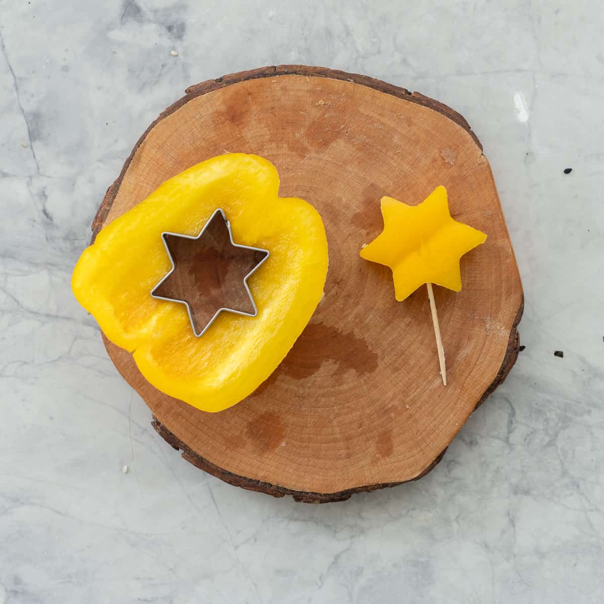 Piece of yellow capsicum with star cookie cutter inside and star cut out with toothpick inside on wooden log ring on benchtop.