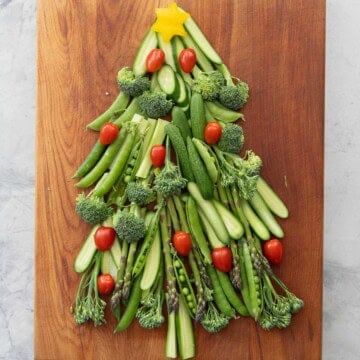 Vegetables laid out on a wooden chopping board as a Christmas tree, cucumber slices, asparagus, broccolini, sweet peas as the branches. Yellow capsicum star cut out on the top and cherry tomatoes as christmas lights.