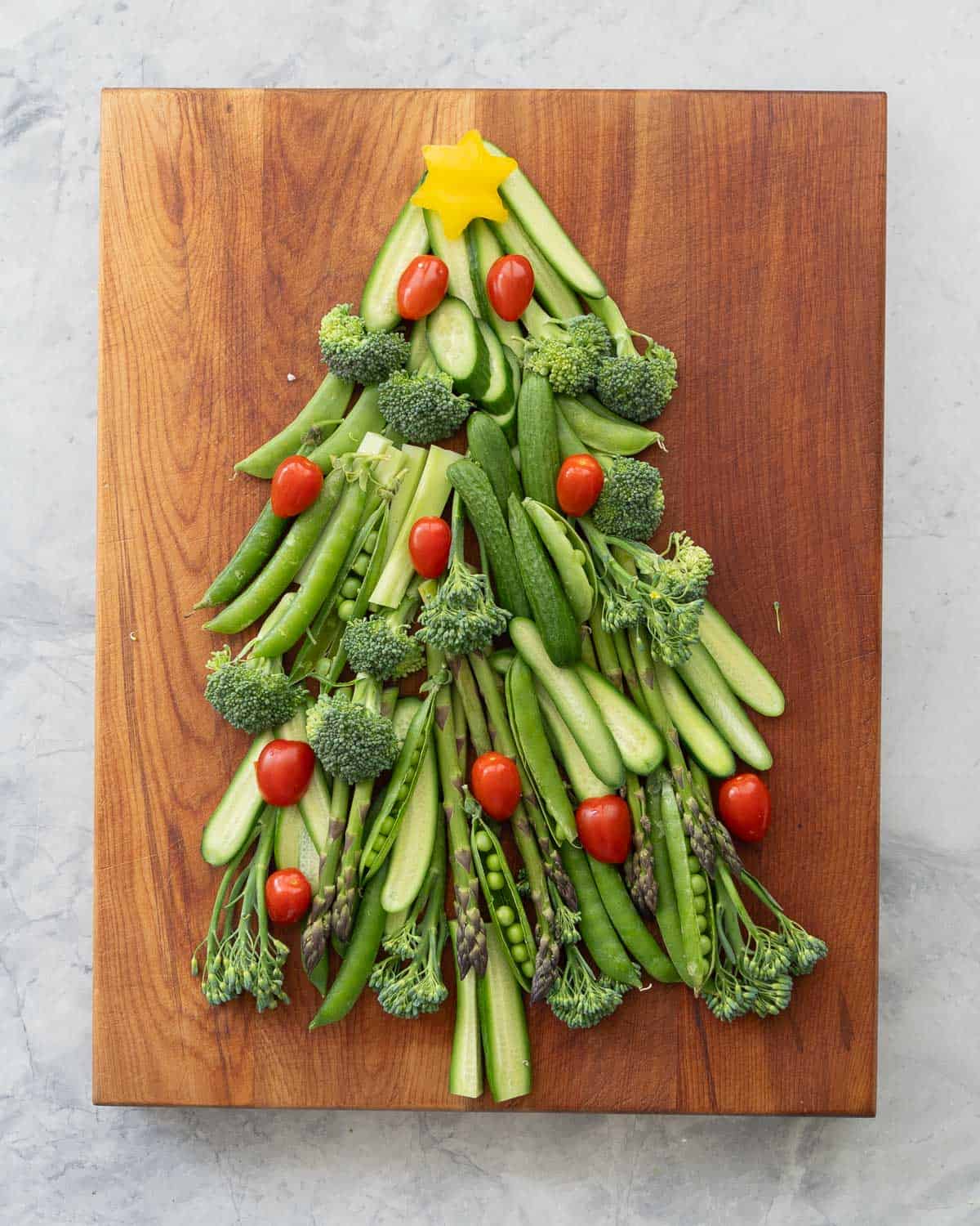 Vegetables laid out on a wooden chopping board as a Christmas tree, cucumber slices, asparagus, broccolini, sweet peas as the branches. Yellow capsicum star cut out on the top and cherry tomatoes as christmas lights.