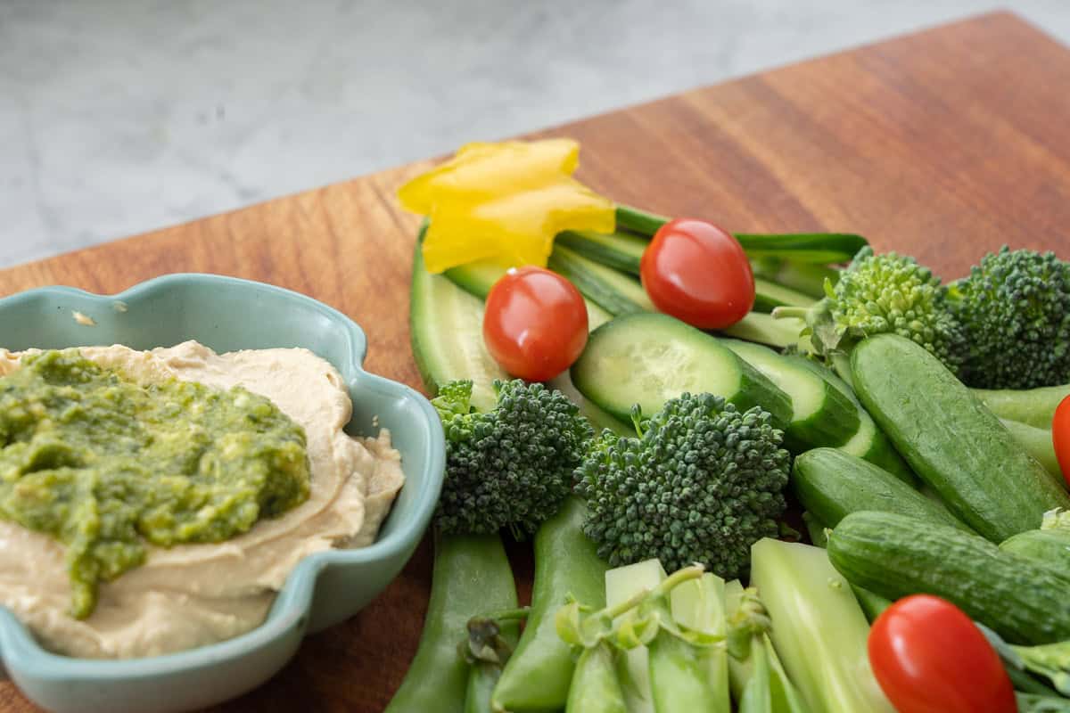 Close up of Vegetables laid out on a wooden chopping board as a Christmas tree, cucumber slices, asparagus, broccolini, sweet peas as the branches. Yellow capsicum star cut out on the top and cherry tomatoes as christmas lights. Small ramekin of hummus and basil pesto on the side.