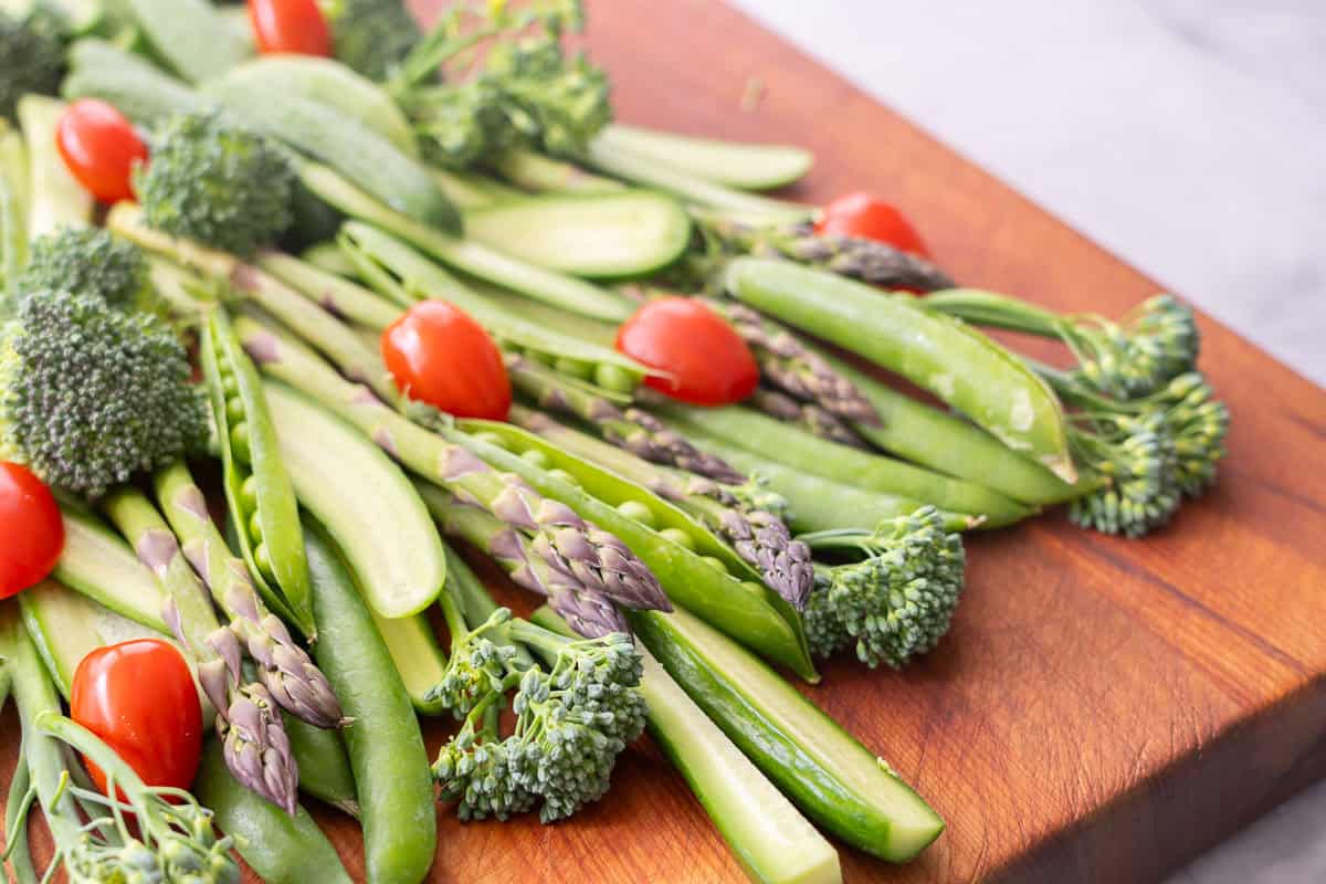 Close up of vegetables laid out on a wooden chopping board as a Christmas tree, cucumber slices, asparagus, broccolini, sweet peas as the branches and cherry tomatoes.