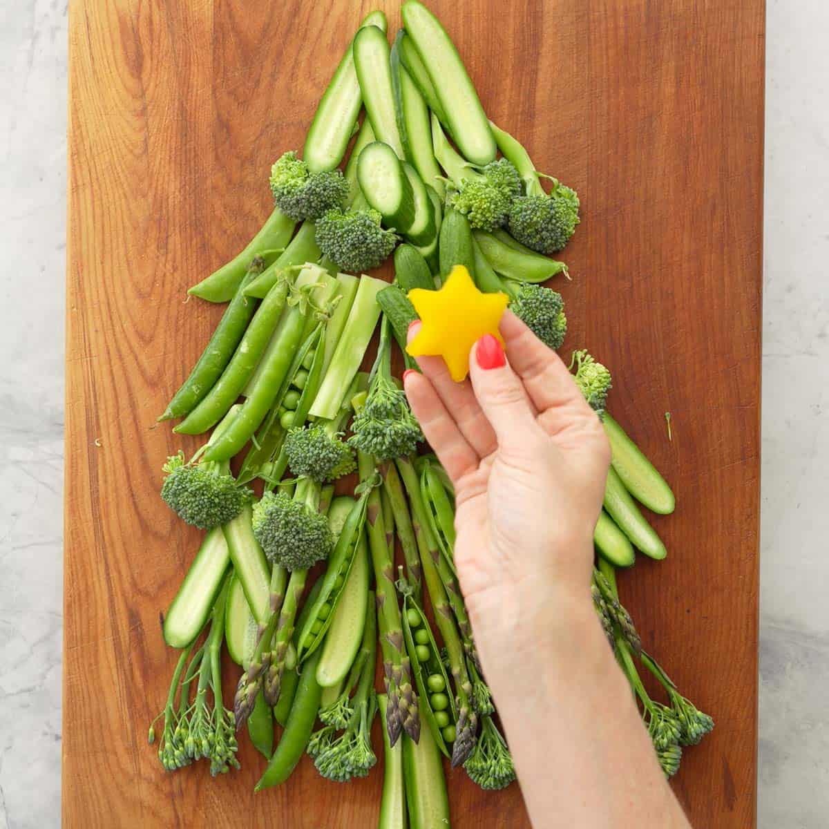 Vegetables laid out on a wooden chopping board as a Christmas tree, cucumber slices, asparagus, broccolini, sweet peas as the branches. Hand holding a yellow capsicum star cut out.