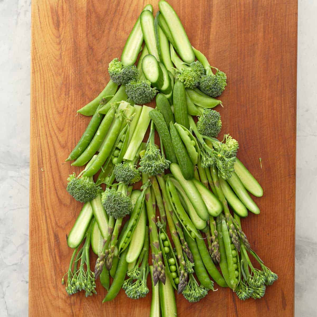 Vegetables laid out on a wooden chopping board as a Christmas tree, cucumber slices, asparagus, broccolini, sweet peas as the branches.