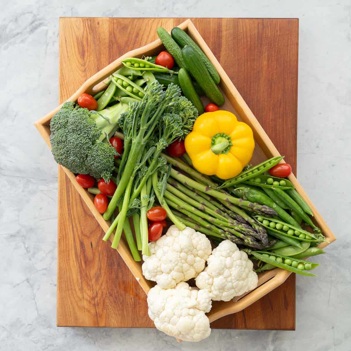 An array of raw Vegetables on a tray siting on wooden chopping board, asparagus, broccolini, yellow capsicum, cucumber, cauliflower cherry tomatoes.