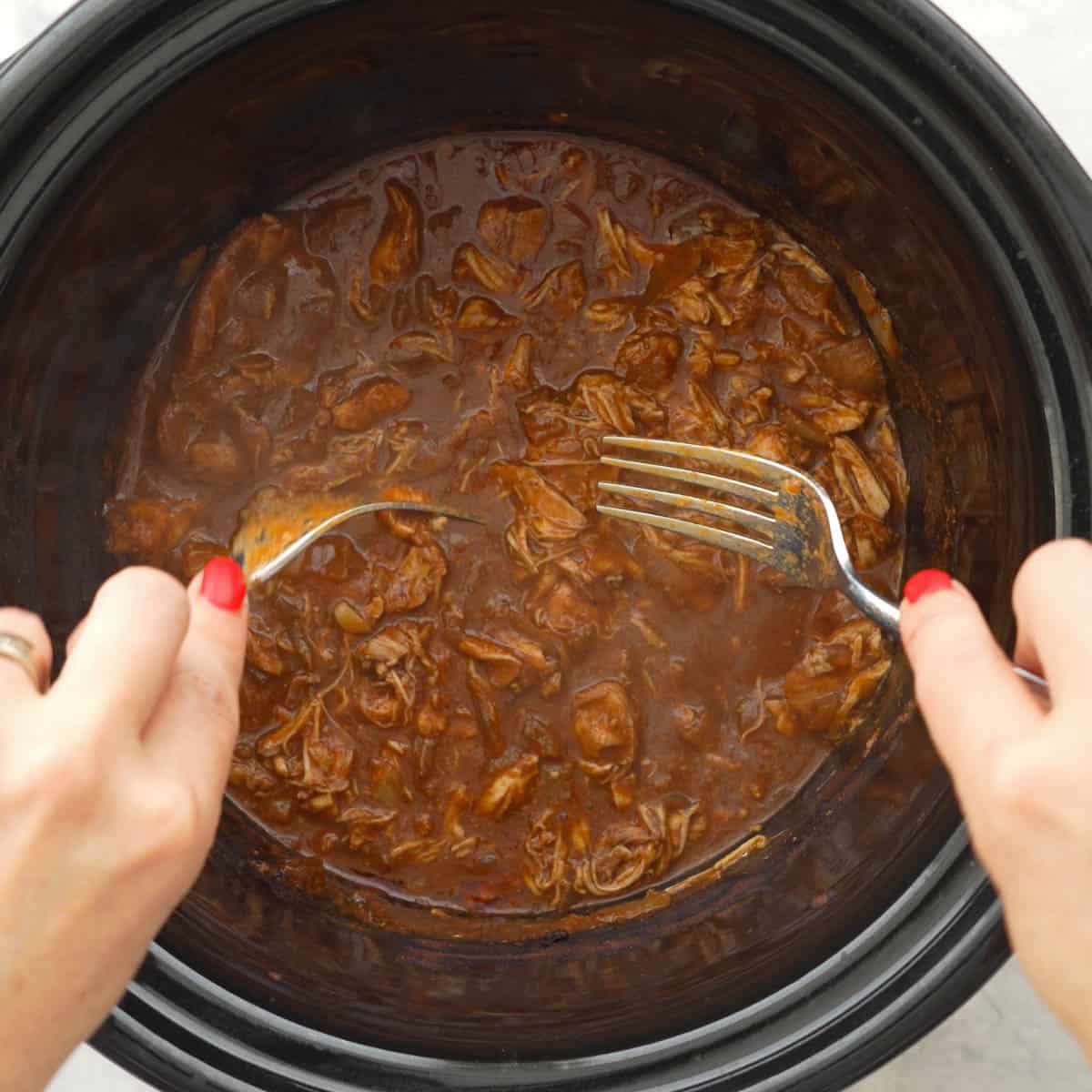 Hands holding two forks in each hands shredding the chicken inside crockpot.