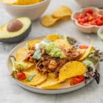 Dinner plate of Crockpot Chicken nachos served on plate with nacho chips, lettuce, cherry tomatoes, mashed avocado and sour cream on top. Small bowls of nacho chips, avocado, lettuce, cherry tomatoes and sour cream on benchtop around dinner plate.