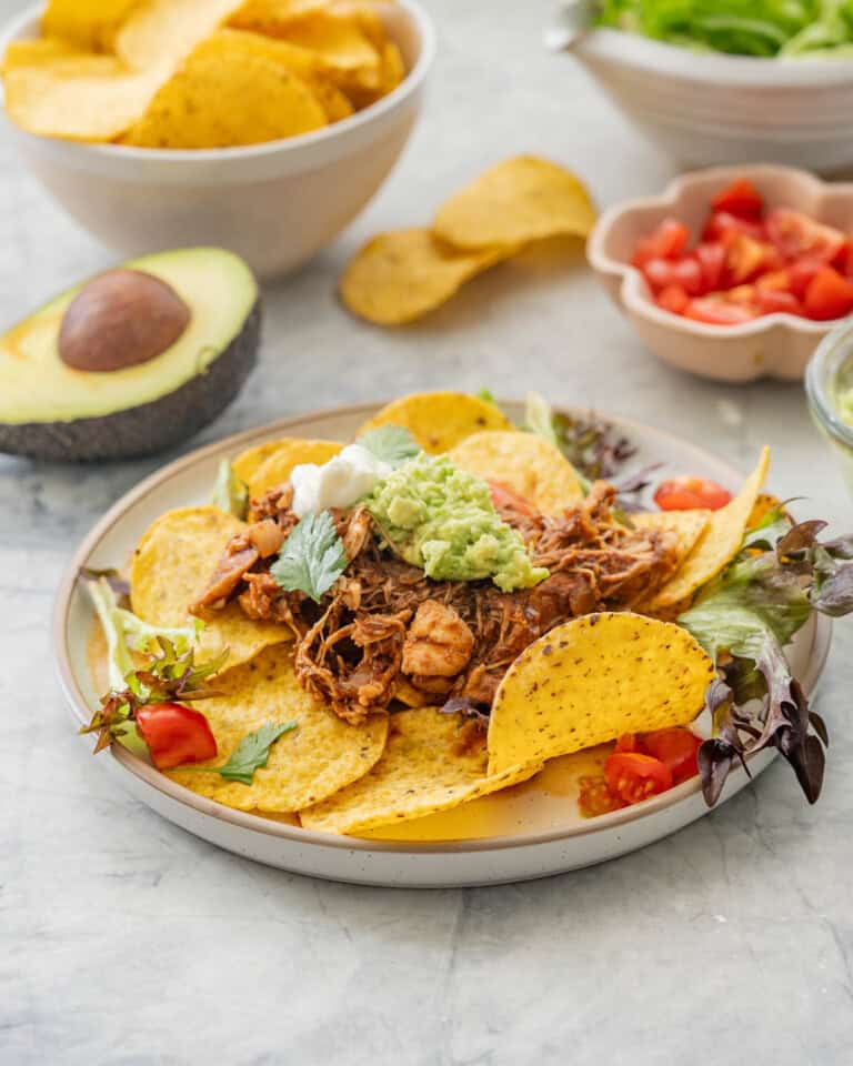 Dinner plate of Crockpot Chicken nachos served on plate with nacho chips, lettuce, cherry tomatoes, mashed avocado and sour cream on top. Small bowls of nacho chips, avocado, lettuce, cherry tomatoes and sour cream on benchtop around dinner plate.