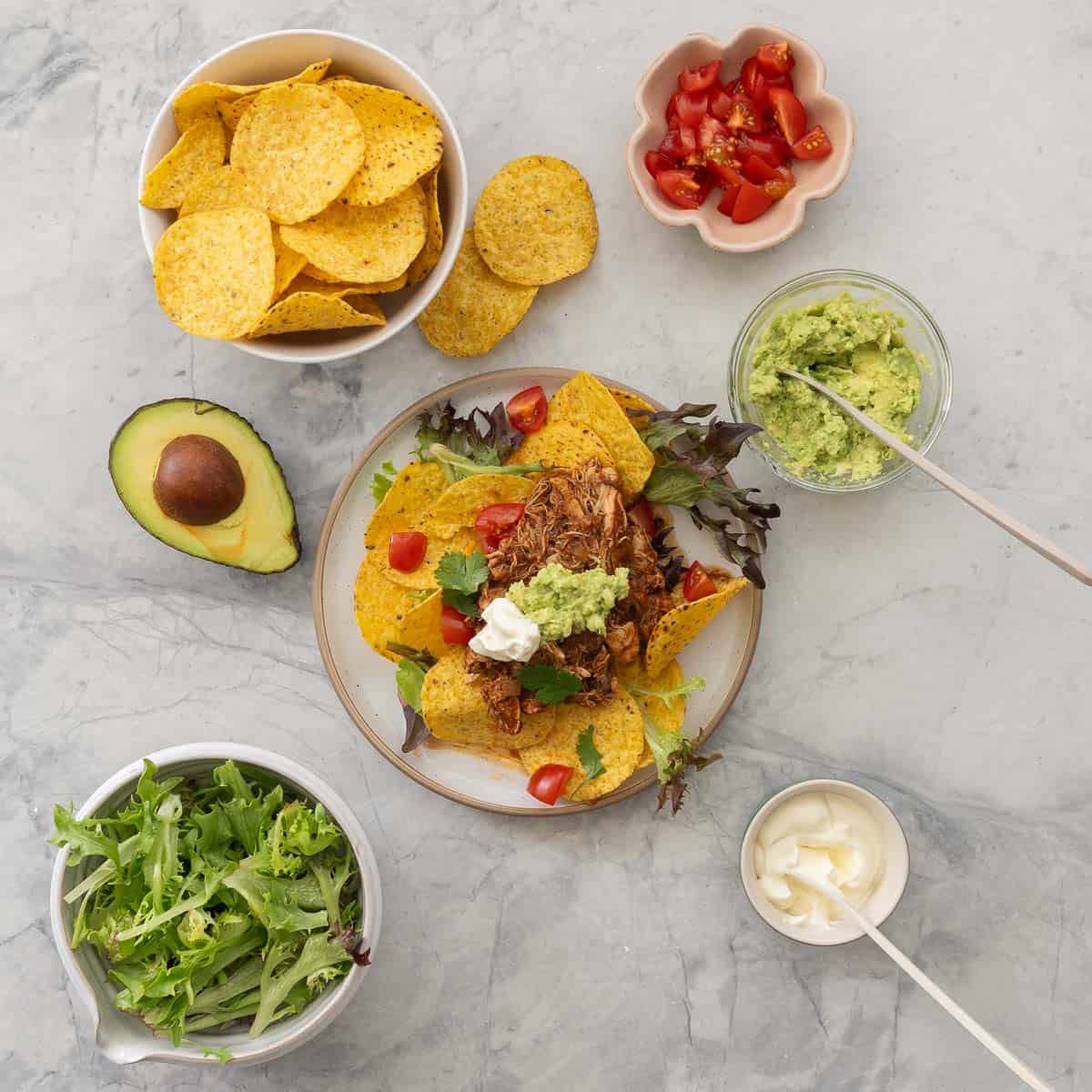 Dinner plate of Crockpot Chicken nachos served on plate with nacho chips, lettuce, cherry tomatoes, mashed avocado and sour cream on top. Small bowls of nacho chips, avocado, lettuce, cherry tomatoes and sour cream on benchtop around dinner plate.