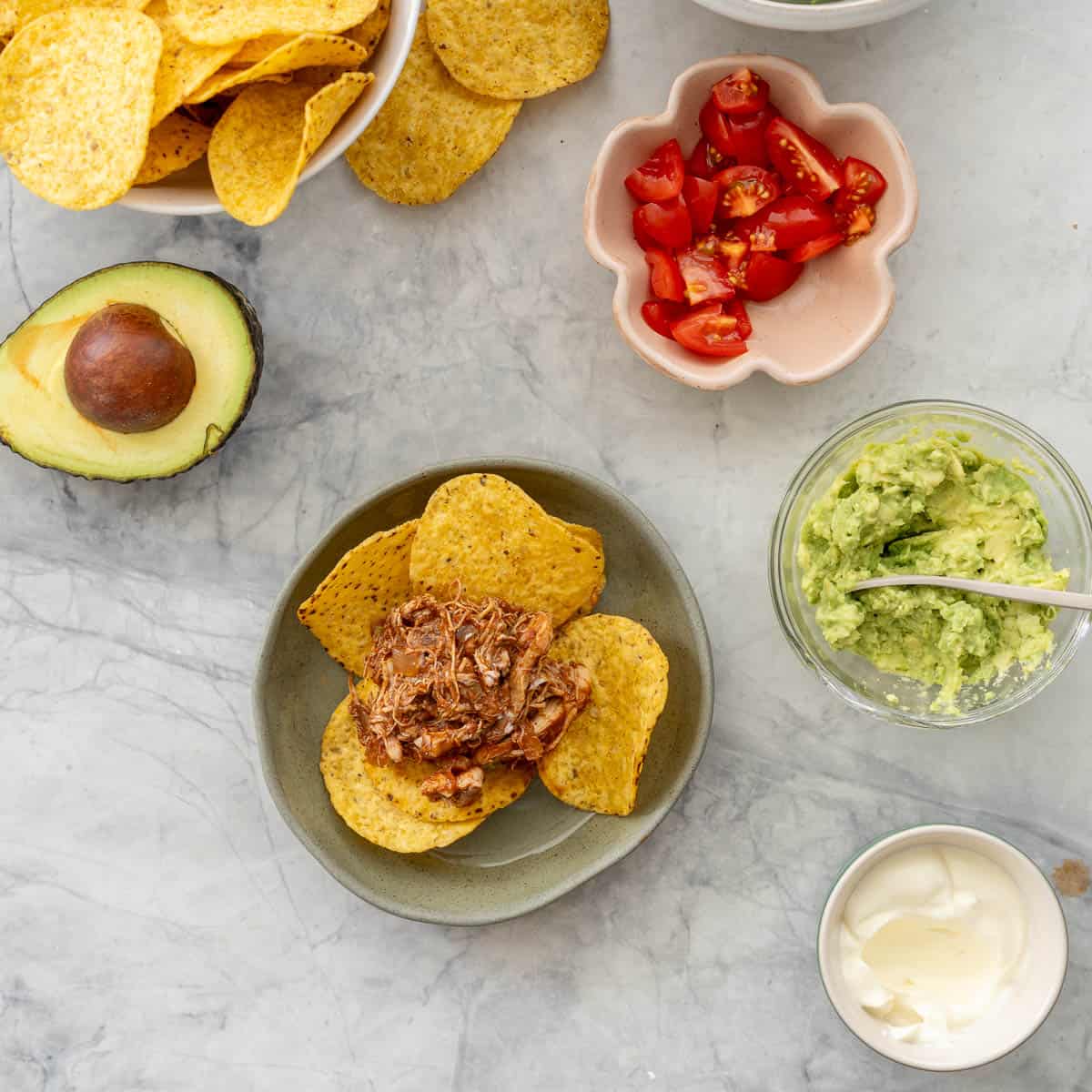 Dinner plate of Crockpot Chicken nachos served on plate with nacho chips. Small bowls of nacho chips, avocado, lettuce, cherry tomatoes and sour cream on benchtop around dinner plate.