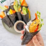 A sushi hand roll filled with colourful fillings being held up above a platter of sushi, with text overlay: Hand roll sushi, the easiest way to make sushi.