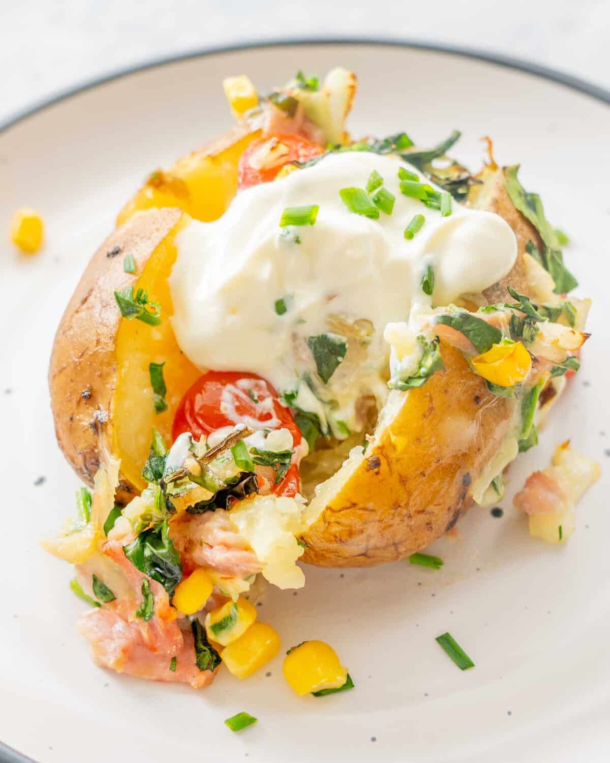 Loaded baked potatoe on a dinner plate with toppings of sweetcorn, cherry tomatoes, spinach, bacon, onion and melted cheese sprinkled on top.