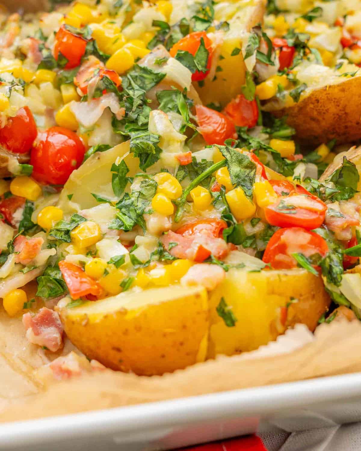 Loaded baked potatoes in baking dish lined with parchment paper with toppings of sweetcorn, cherry tomatoes, spinach, bacon, onion and melted cheese sprinkled on top of cooked potato.