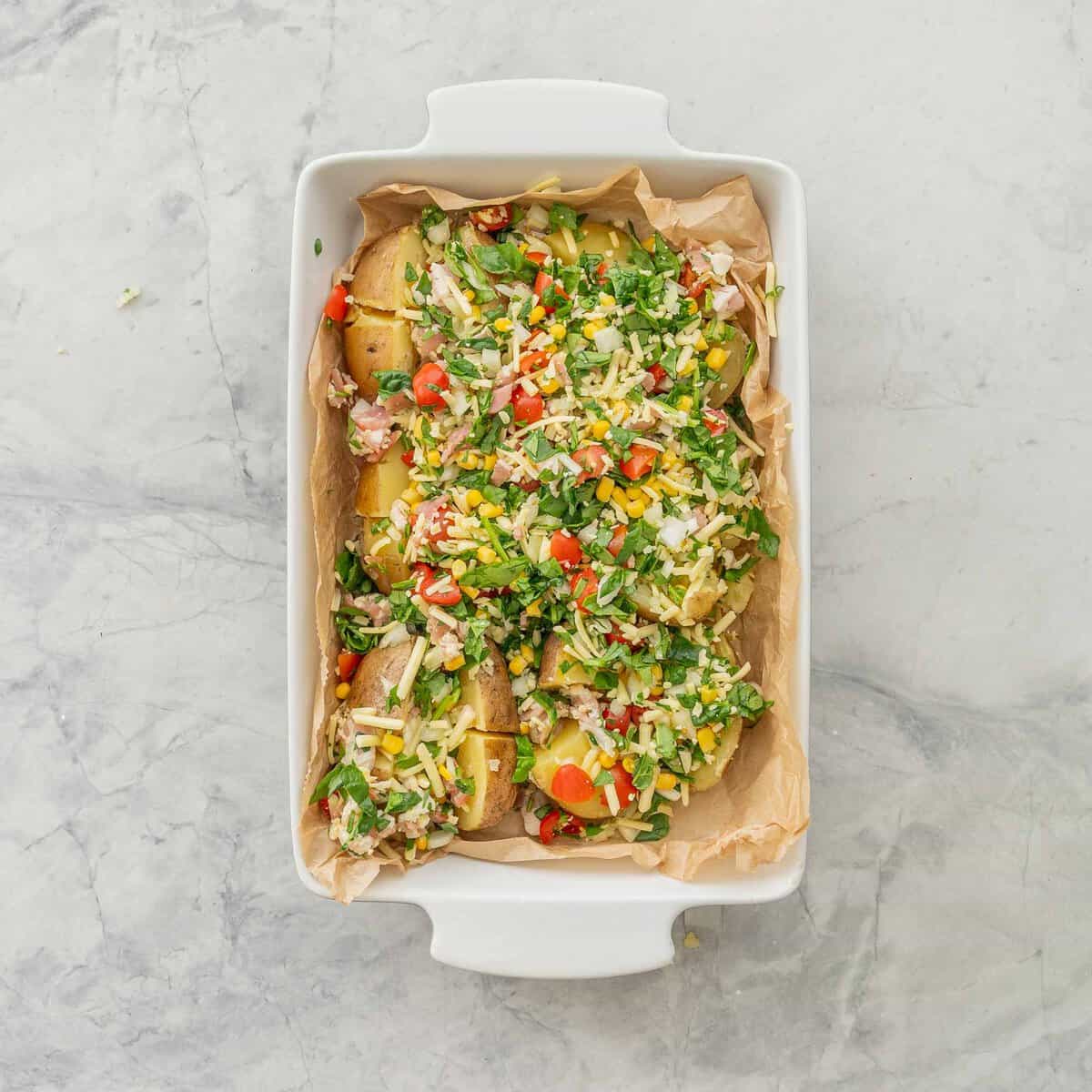 Loaded baked potatoes in baking dish lined with parchment paper with toppings of sweetcorn, cherry tomatoes, spinach, bacon, onion and cheese ready to go into oven.