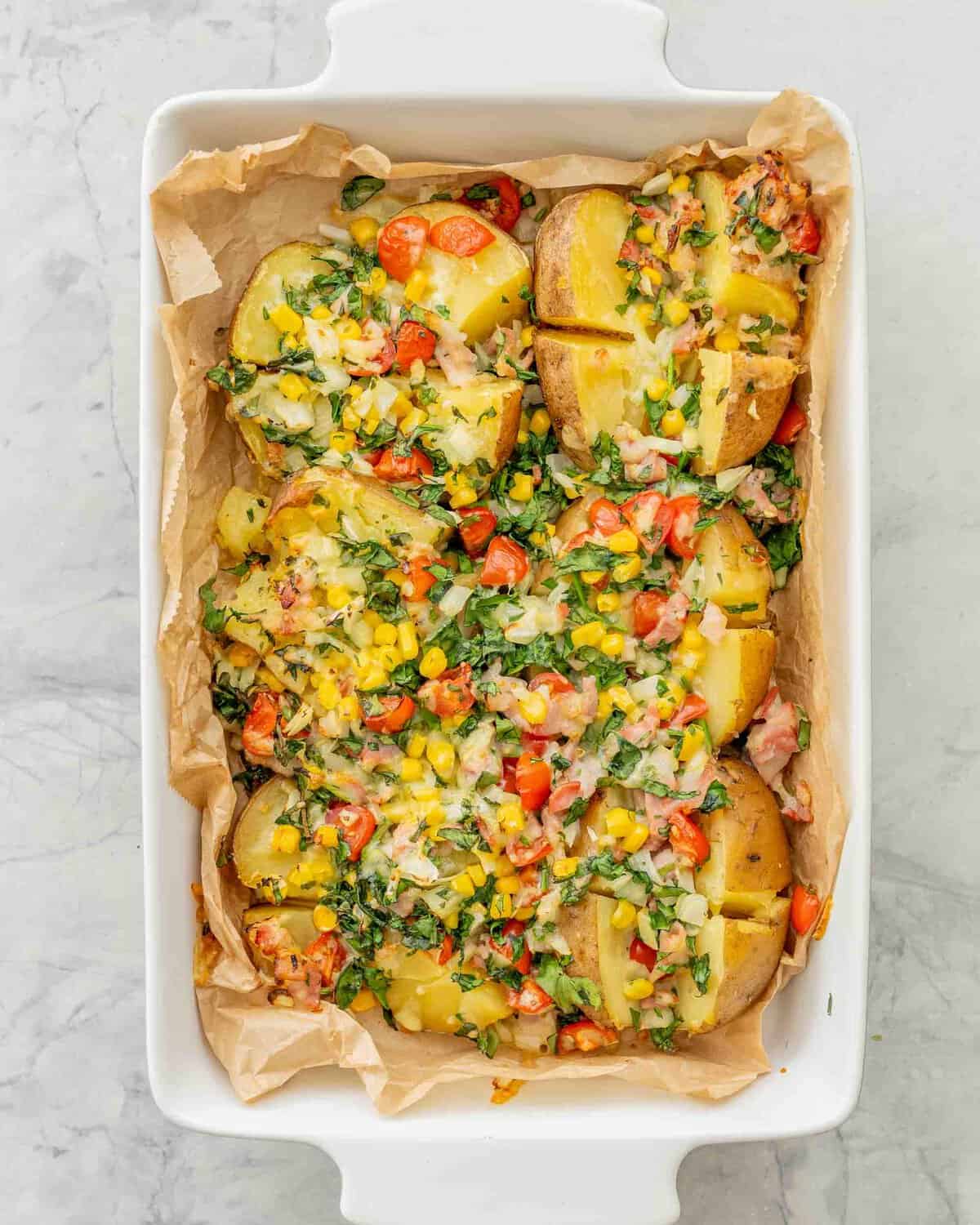 Loaded baked potatoes in baking dish lined with parchment paper with toppings of sweetcorn, cherry tomatoes, spinach, bacon, onion and melted cheese sprinkled on top of cooked potato.