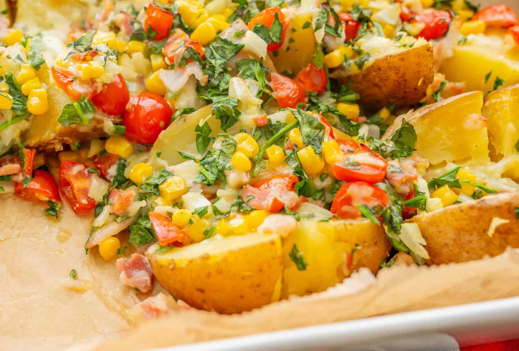 Loaded baked potatoes in baking dish lined with parchment paper with toppings of sweetcorn, cherry tomatoes, spinach, bacon, onion and melted cheese sprinkled on top of cooked potato.