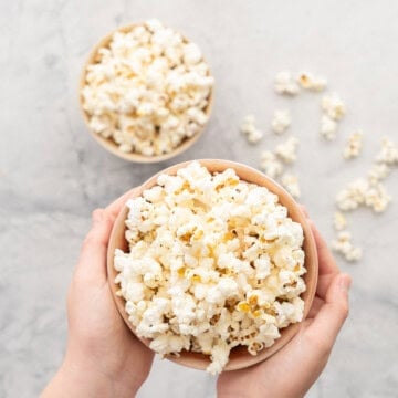 A bowl of popcorn being held but children's hands being held above a bench top which has more bowls of popcorn on it.