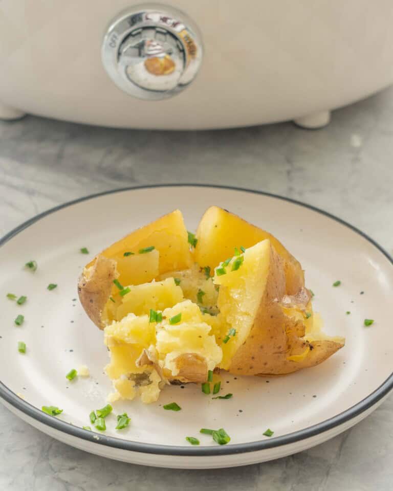 Slow cooker baked potato on a dinner plate cut in half with melted butter inside and sprinkled with chives.