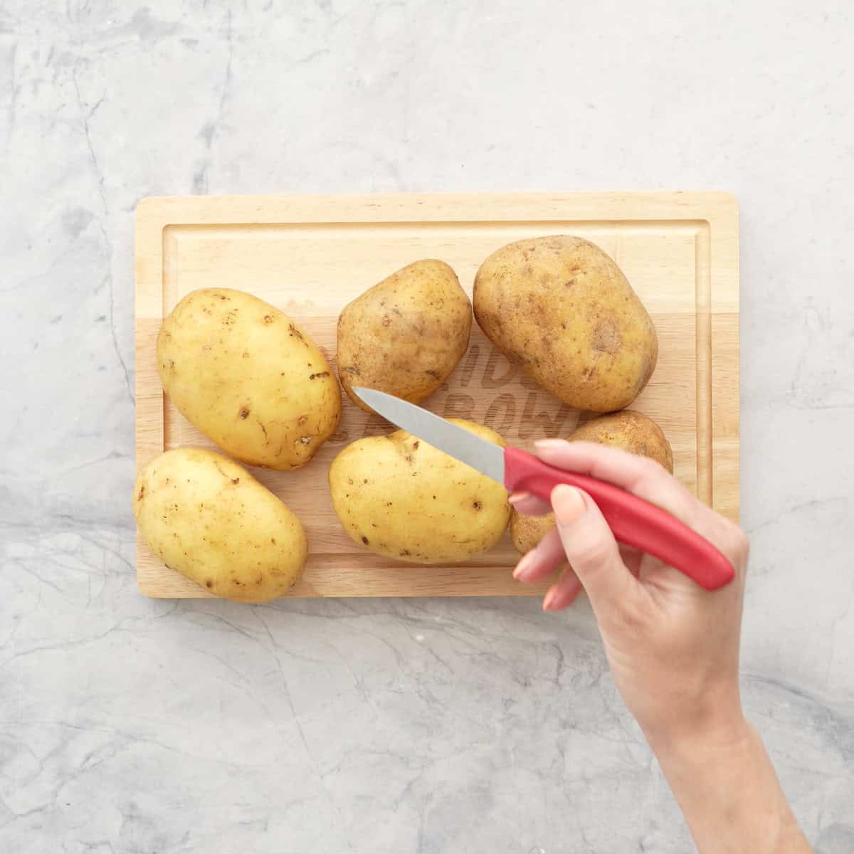 Raw Potatoes on a wooden chopping board with hand holding a sharp knife.