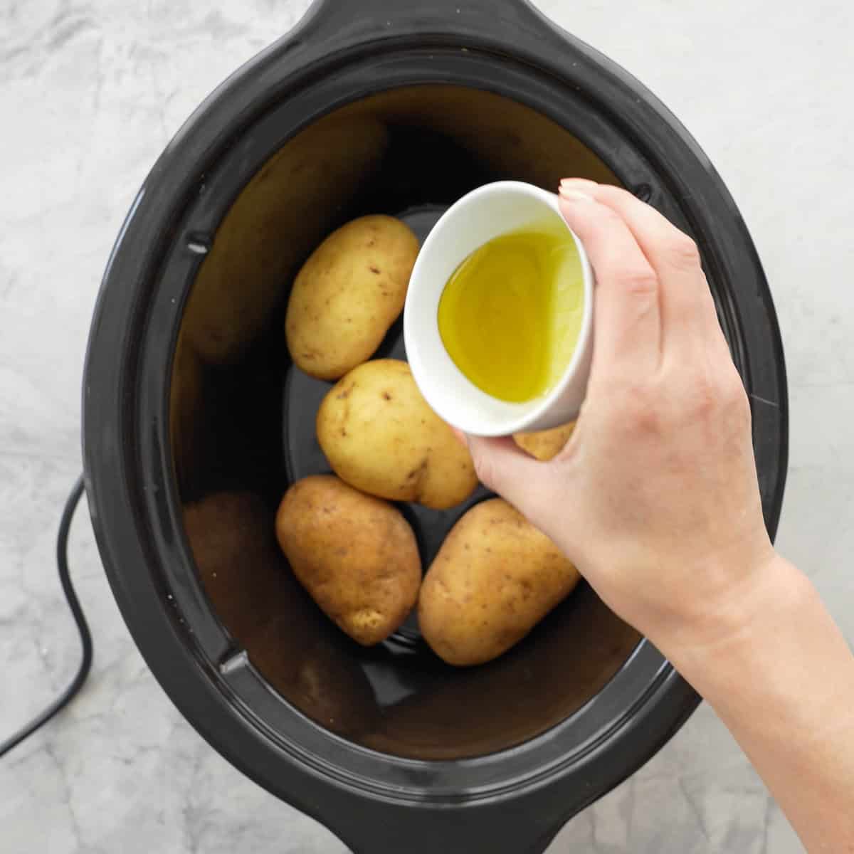 Hand holding a ceramic dish with olive oil being poured on top of raw potatoes inside slow cooker.