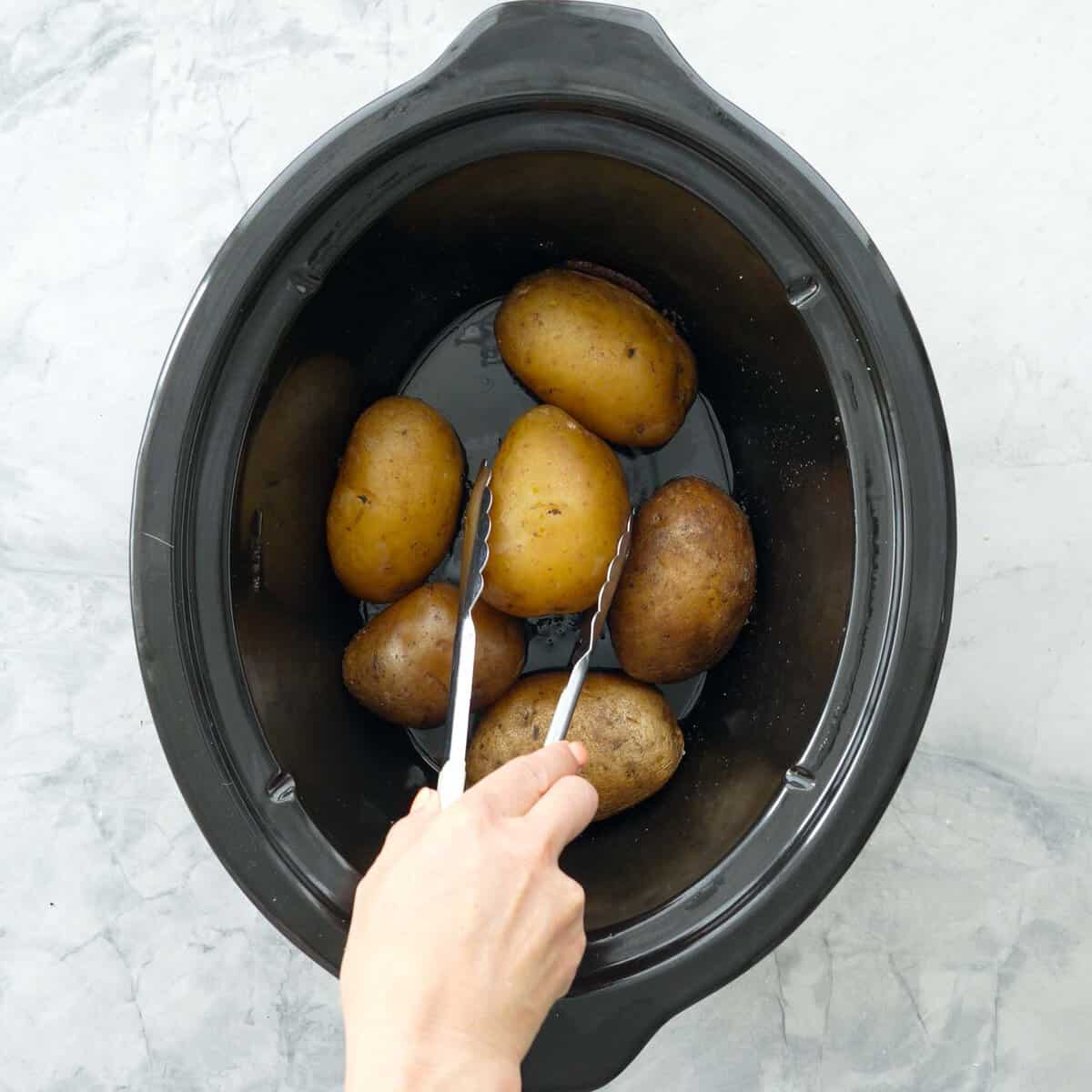 Hand holding a a pair of tongs turning over a potato inside the slow cooker.