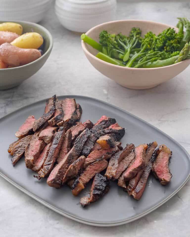 Marinated steak on a big plate sliced into thin pieces showing a medium cook inside, asparagus and broccolini in a bowl and seasoned potatoes on a bowl on bench top.