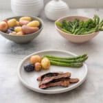 Dinner plate with marinated steak, potatoes and asparagus, bowl of broccolini and asparagus in a bowl and seasons potatoes in bowl on benchtop.