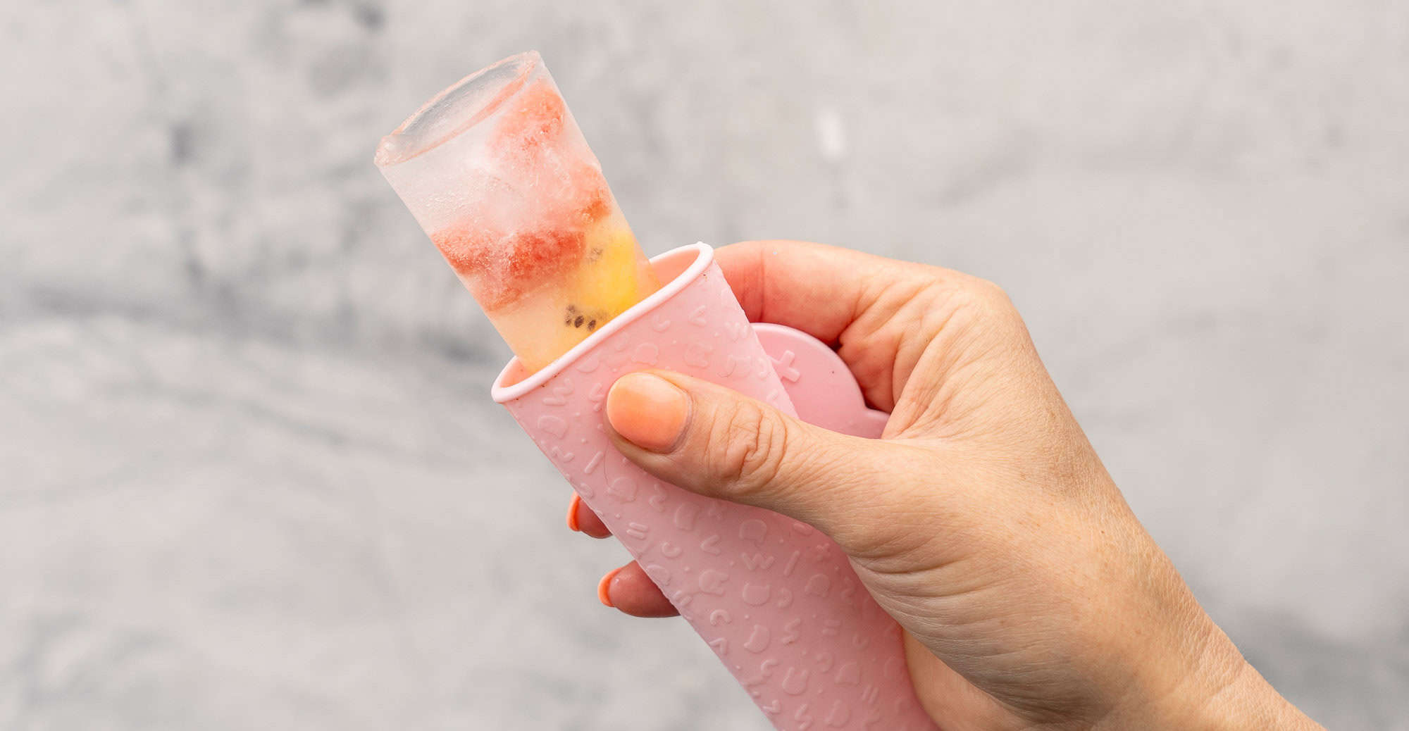 Hand holding a pink Popsicle holder with water and fruit ice block showing out of the top.