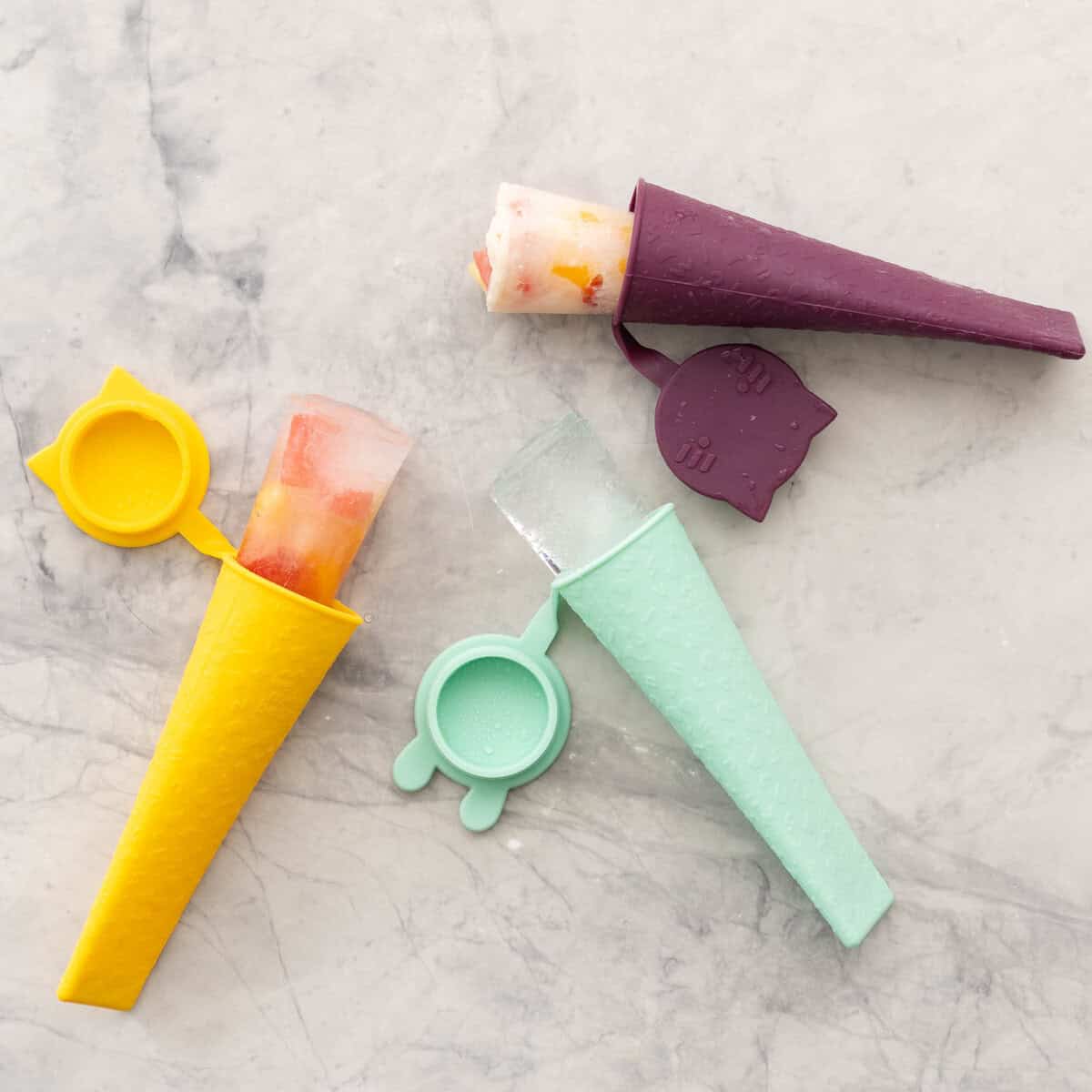 Three popsicles holders laid on benchtop, one with frozen water and fruit inside, another with frozen milk and fruit and one with ice.