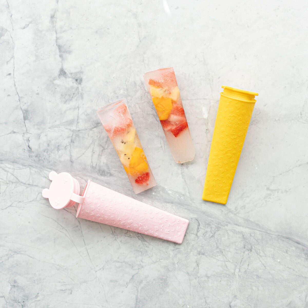 Two water and fruit ice blocks on bench top with two silicone popsicle holders.