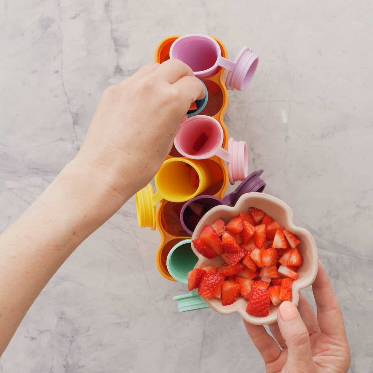 Hand holding a ceramic bowl with cut up strawberries inside with other hand placing them into silicone holders.